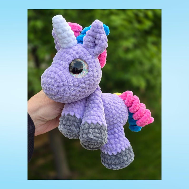 Peut inclure: Une peluche licorne crochet&eacute;e &agrave; la main, dans des tons de violet, de gris et d'accents color&eacute;s. La licorne a une corne blanche, une crini&egrave;re rose et bleue, et une queue boucl&eacute;e avec des sections roses et bleues. La peluche a de grands yeux brillants.