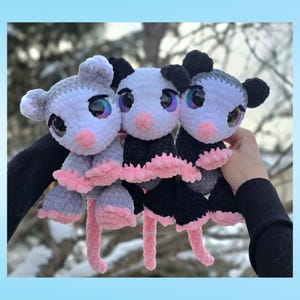 Pu&ograve; includere: Tre peluche di opossum all'uncinetto fatti a mano. Gli opossum hanno grandi occhi iridescenti, nasi rosa e corpi rosa e grigi o neri e bianchi. I peluche sono tenuti contro uno sfondo sfocato all'aperto.