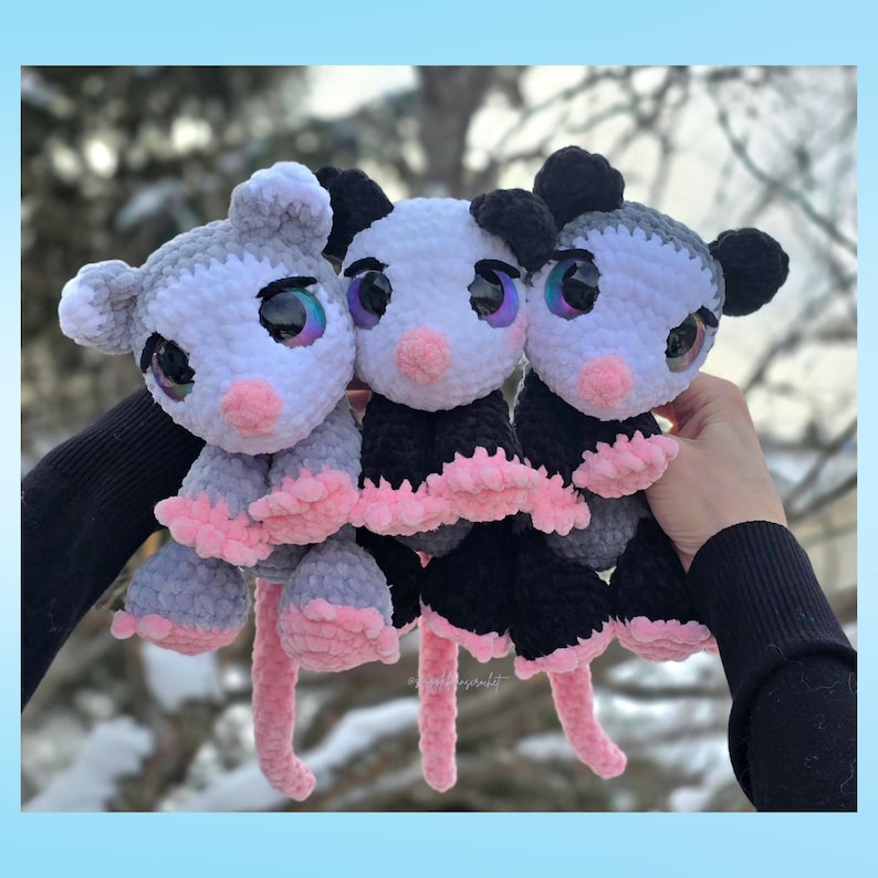 Pu&ograve; includere: Tre peluche di opossum all'uncinetto fatti a mano. Gli opossum hanno grandi occhi iridescenti, nasi rosa e corpi rosa e grigi o neri e bianchi. I peluche sono tenuti contro uno sfondo sfocato all'aperto.