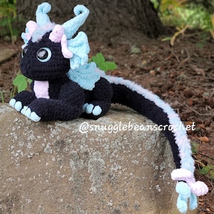 May include: A black crocheted dragon with blue and pink accents. The dragon is sitting on a rock with its tail curled around the rock. The dragon has large, black eyes and a pink nose. The dragon's wings are blue and pink, and its tail is long and thin. The dragon is a handmade crocheted toy.