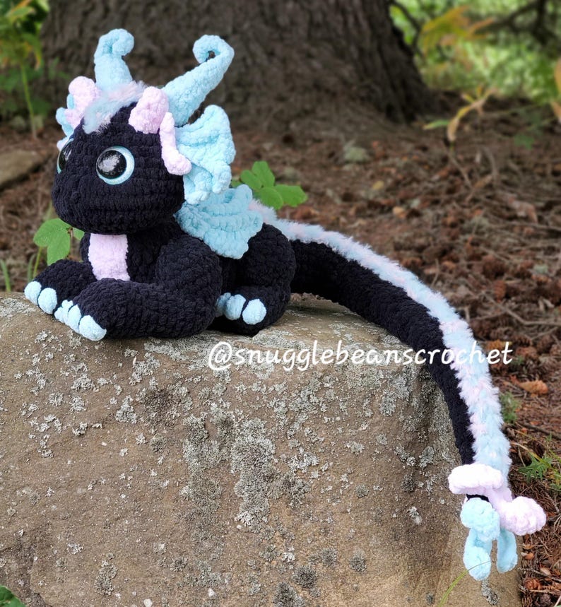 May include: A black crocheted dragon with blue and pink accents. The dragon is sitting on a rock with its tail curled around the rock. The dragon has large, black eyes and a pink nose. The dragon's wings are blue and pink, and its tail is long and thin. The dragon is a handmade crocheted toy.