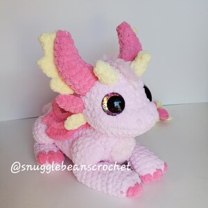 May include: A pink and yellow crocheted dragon plush toy with large, glittery eyes. The dragon is sitting on a white surface. @snugglebeanscrochet