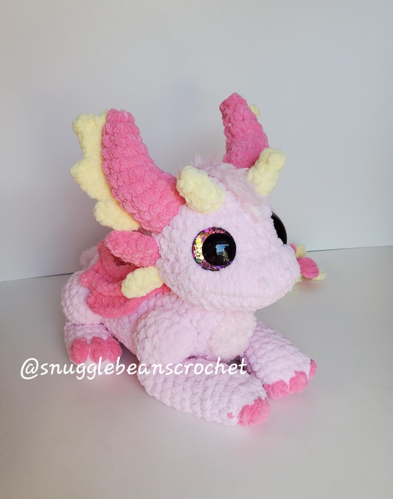 May include: A pink and yellow crocheted dragon plush toy with large, glittery eyes. The dragon is sitting on a white surface. @snugglebeanscrochet