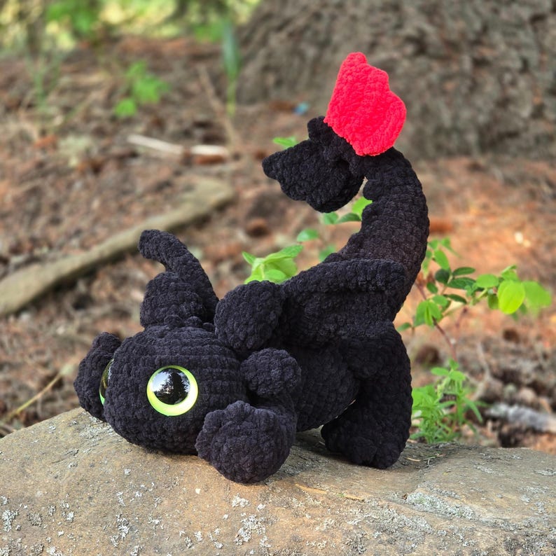 Pode incluir: Um brinquedo de pel&uacute;cia de drag&atilde;o preto feito &agrave; m&atilde;o com grandes olhos verdes e uma crista vermelha. O drag&atilde;o est&aacute; deitado em uma rocha, com o corpo curvado e a cauda levantada. A pel&uacute;cia &eacute; feita de fio macio e texturizado.