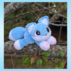 Puede incluir: Un peluche de zorro hecho a mano de ganchillo azul y blanco con ojos grandes y expresivos y almohadillas rosas. El peluche est&aacute; tumbado en una rama de &aacute;rbol, mostrando su textura suave y su detallada artesan&iacute;a. El zorro tiene una apariencia linda y amigable.