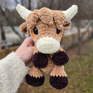 May include: A handmade crochet Highland cow plushie. The toy has a light brown body, dark brown hooves, and a white muzzle and horns. It has large, orange eyes. The plushie is held in a person's hand.