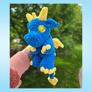 May include: A blue and yellow crocheted dragon plush toy. The dragon has yellow horns, wings, and feet. The eyes are yellow with gold glitter. The plush is held in a person's hand.