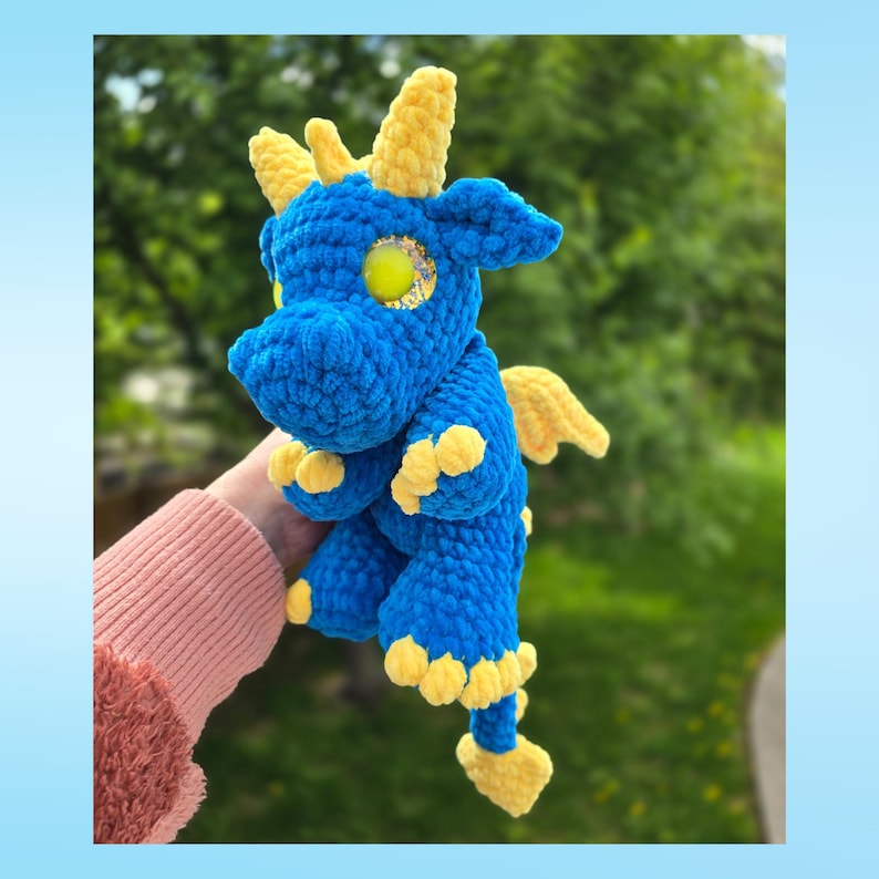 May include: A blue and yellow crocheted dragon plush toy. The dragon has yellow horns, wings, and feet. The eyes are yellow with gold glitter. The plush is held in a person's hand.