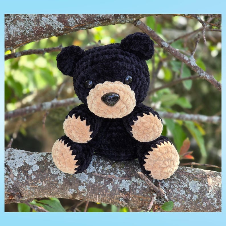 May include: A handmade black bear plush toy with tan accents on its snout, paws, and ears. The bear has black eyes and a black nose. It is sitting on a tree branch with a blurred green background.