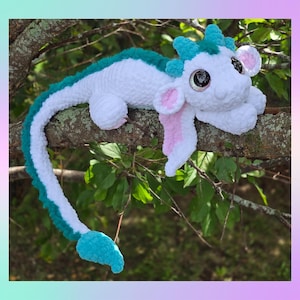 May include: A handmade white and teal crochet dragon plush toy with large, glittery eyes, pink inner ears, and a long tail. The dragon is lying on a tree branch, showcasing its soft, textured design. The dragon's horns and spine are teal.
