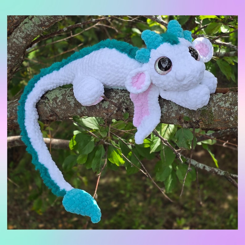 May include: A handmade white and teal crochet dragon plush toy with large, glittery eyes, pink inner ears, and a long tail. The dragon is lying on a tree branch, showcasing its soft, textured design. The dragon's horns and spine are teal.