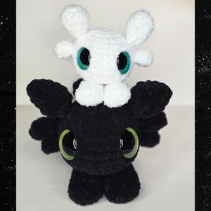Puede incluir: Un peluche hecho a mano con un drag&oacute;n negro con grandes ojos verdes y una criatura blanca con ojos verdes encaramada encima. Los juguetes est&aacute;n hechos de un material suave y texturizado.