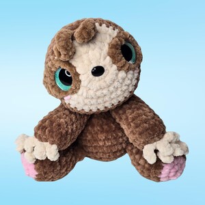 Pode incluir: Um brinquedo de pel&uacute;cia de croch&ecirc; feito &agrave; m&atilde;o em forma de um pequeno animal. O brinquedo apresenta um corpo marrom, um rosto creme e olhos grandes com detalhes em turquesa. As patas t&ecirc;m solas rosa. A pel&uacute;cia &eacute; macia e fofinha.
