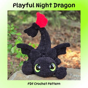 Pode incluir: Um dragão preto de crochê com olhos verdes grandes e uma barbatana vermelha. O dragão está sentado em uma rocha com um fundo verde desfocado. O texto "Playful Night Dragon" e "PDF Crochet Pattern" são visíveis.