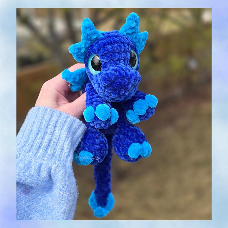 May include: A handmade blue crochet dragon with large, round eyes and small wings. The dragon has a textured, plush appearance with lighter blue accents on its claws, horns, and tail. The dragon is a whimsical, fantasy-themed item.