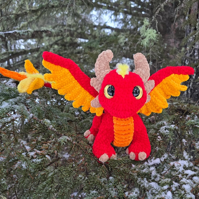 May include: A crocheted red dragon with orange wings and yellow accents. The dragon is sitting on a snowy branch of a pine tree.