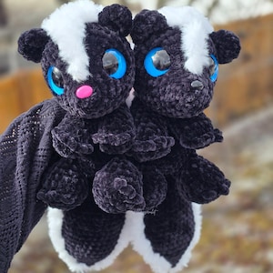 May include: Two handmade crochet skunk plush toys. The skunks are black with white stripes and accents, large blue eyes, and pink noses. They are soft and cuddly, ideal as a child's toy or a unique gift.