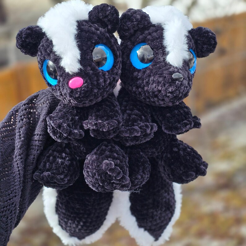 May include: Two handmade crochet skunk plush toys. The skunks are black with white stripes and accents, large blue eyes, and pink noses. They are soft and cuddly, ideal as a child's toy or a unique gift.