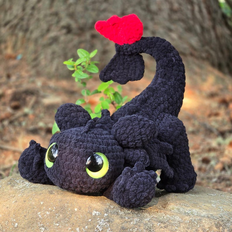 Pode incluir: Um peluche de drag&atilde;o preto de croch&ecirc; feito &agrave; m&atilde;o com olhos grandes e expressivos e uma barbatana vermelha. O drag&atilde;o est&aacute; deitado numa rocha, com uma planta verde ao fundo. O peluche &eacute; feito de fio macio e texturizado.