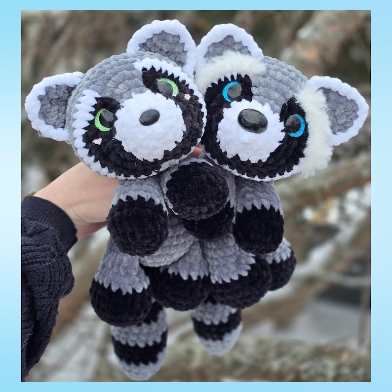 May include: Two handmade crochet raccoon plush toys. The raccoons have gray and white fur with black markings around their eyes and on their paws. One raccoon has green eyes, and the other has blue eyes. The plush toys are soft and cuddly.