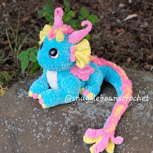 May include: A crocheted plush dragon toy with a blue body, pink and yellow accents, and large, sparkly eyes. The dragon has a long, curled tail and is sitting on a rock. The text "@snugglebeanscrochet" is visible in the image.
