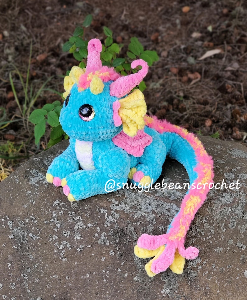 May include: A crocheted plush dragon toy with a blue body, pink and yellow accents, and large, sparkly eyes. The dragon has a long, curled tail and is sitting on a rock. The text "@snugglebeanscrochet" is visible in the image.