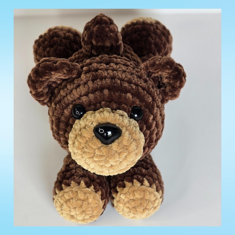 May include: A handmade brown crochet bear with a light brown muzzle and black eyes. The bear has a soft, plush texture and is designed to sit upright. The bear's ears and head are rounded, and the nose is a glossy black.