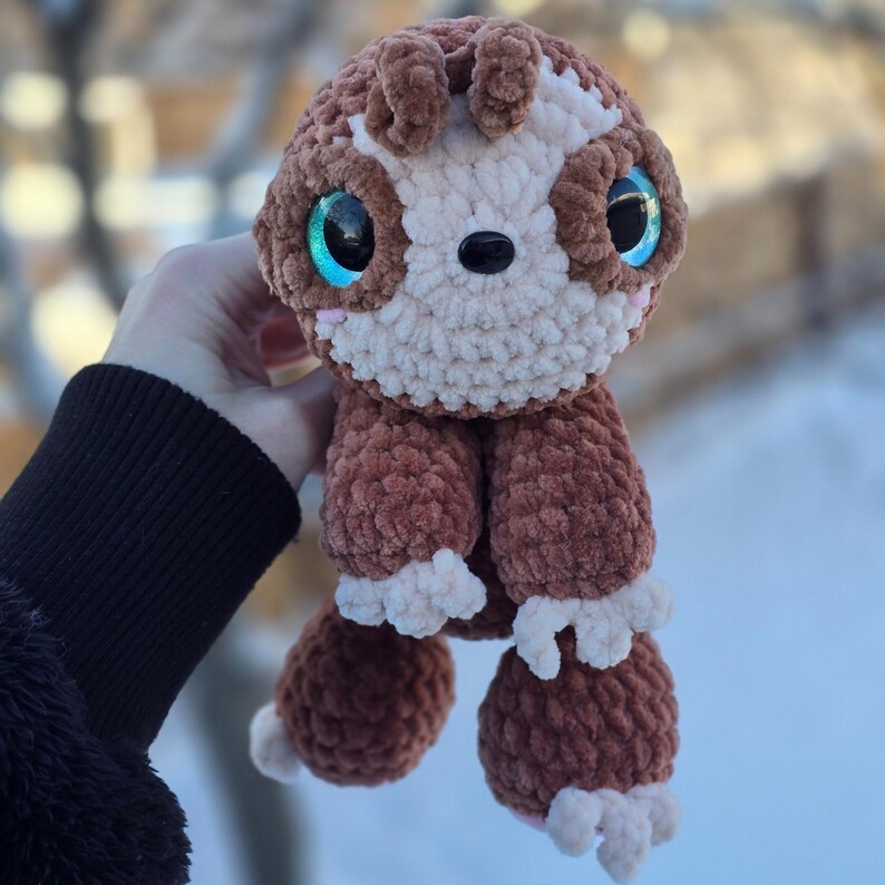 Pode incluir: Um animal de croch&ecirc; marrom e creme feito &agrave; m&atilde;o com grandes olhos azuis e um nariz preto. O brinquedo de pel&uacute;cia tem uma cabe&ccedil;a redonda, orelhas pequenas e quatro pernas curtas com patas cor creme. Fundo desfocado.