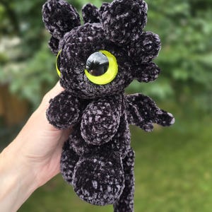 Puede incluir: Un drag&oacute;n de peluche negro hecho a mano con grandes ojos amarillos y peque&ntilde;as alas. El drag&oacute;n tiene una cola roja y est&aacute; hecho de un material suave y texturizado. El peluche se sostiene frente a un fondo verde borroso.