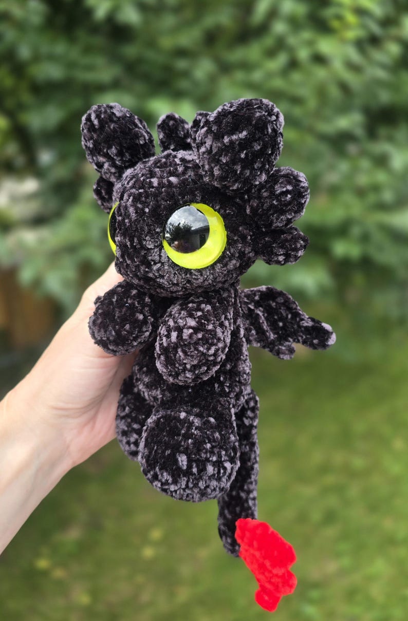 Puede incluir: Un drag&oacute;n de peluche negro hecho a mano con grandes ojos amarillos y peque&ntilde;as alas. El drag&oacute;n tiene una cola roja y est&aacute; hecho de un material suave y texturizado. El peluche se sostiene frente a un fondo verde borroso.