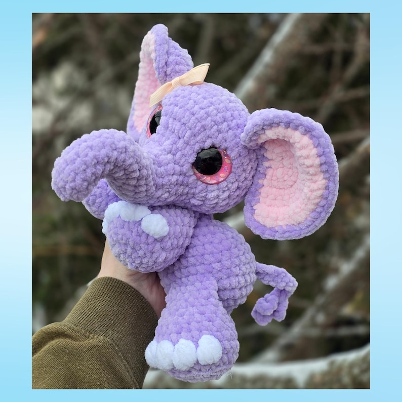 K&ouml;nnte beinhalten: Ein handgefertigtes, pl&uuml;schiges Lavendel-Elefanten-Spielzeug mit gro&szlig;en, rosa umrandeten Augen und einer kleinen Pfirsichschleife. Der Elefant hat gro&szlig;e Ohren mit rosa Innendetails und wei&szlig;en Akzenten an den F&uuml;&szlig;en. Das Stofftier ist mit einem abgerundeten K&ouml;rper und R&uuml;ssel gestaltet.