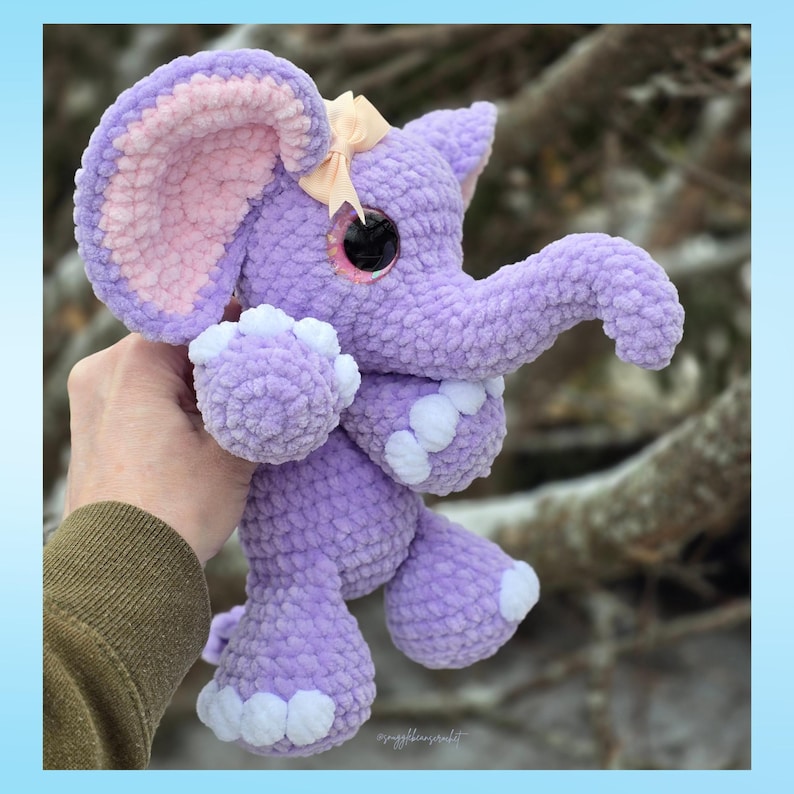K&ouml;nnte beinhalten: Ein handgefertigtes, pl&uuml;schiges lila Elefanten-Spielzeug mit gro&szlig;en Ohren, einem R&uuml;ssel und kleinen wei&szlig;en Akzenten an den F&uuml;&szlig;en. Der Elefant hat eine hellrosa Innenseite an den Ohren und eine kleine Schleife auf dem Kopf. Das Spielzeug wird von einer Person gehalten.