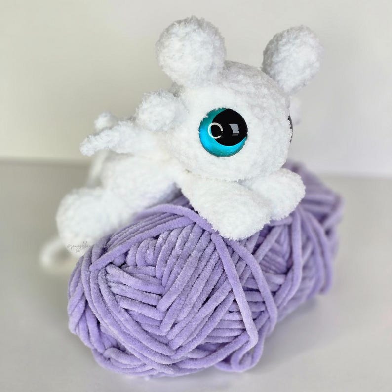 Puede incluir: Un juguete de peluche blanco con forma de drag&oacute;n y grandes ojos azules descansa sobre una bola de hilo lavanda. El drag&oacute;n tiene peque&ntilde;as alas y orejas. El hilo tiene una textura suave y aterciopelada.