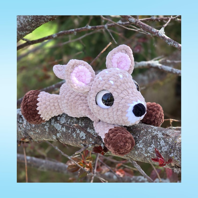 May include: A crocheted fawn toy in light brown with white spots, pink inner ears, and dark brown hooves. The fawn has large eyes and a black nose. It rests on a tree branch, highlighting its soft texture. The toy is suitable for children.