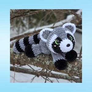 May include: A handmade plush raccoon toy in gray, black, and white, with green eyes, resting on a tree branch. The raccoon has a striped tail and is made of soft, textured material. The background shows a snowy, outdoor scene.