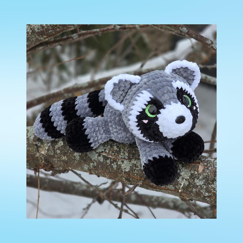 May include: A handmade plush raccoon toy in gray, black, and white, with green eyes, resting on a tree branch. The raccoon has a striped tail and is made of soft, textured material. The background shows a snowy, outdoor scene.