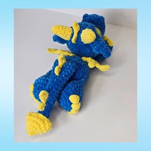 May include: A handmade plush toy in the shape of a dragon. The dragon is primarily blue with yellow accents on its horns, wings, and feet. The plush is made of a soft, textured material.