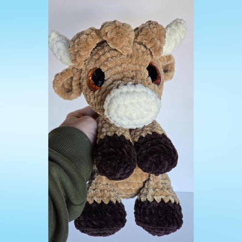 May include: A crocheted cow toy in shades of brown, cream, and dark brown. The cow features large, shiny eyes, white horns, and a soft, textured body. The hooves are dark brown. A cuddly toy for children.