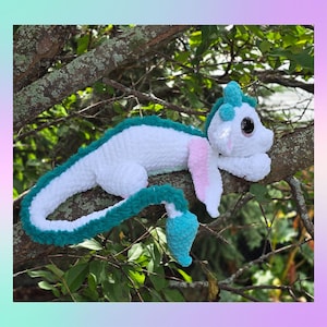 May include: A handmade crochet dragon plush toy in white, teal, and pink, resting on a tree branch. The dragon has large, black eyes and a long, curved tail. The plush is soft and fluffy, with a whimsical design.