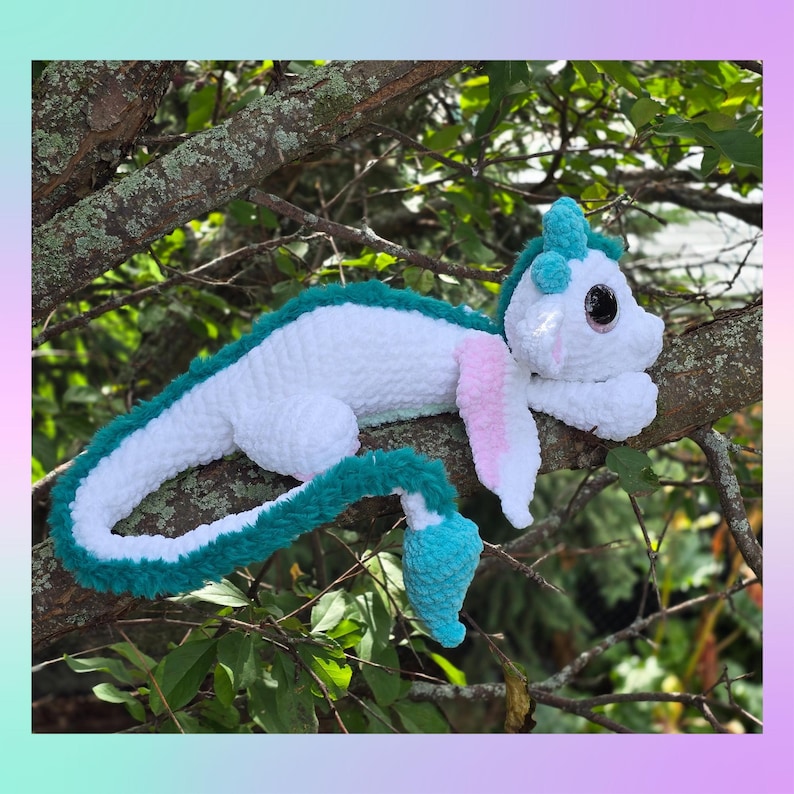 May include: A handmade crochet dragon plush toy in white, teal, and pink, resting on a tree branch. The dragon has large, black eyes and a long, curved tail. The plush is soft and fluffy, with a whimsical design.