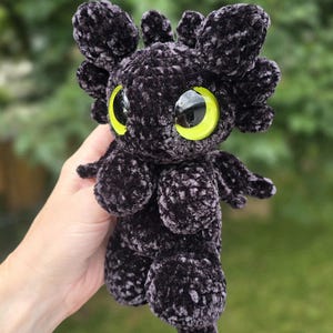 Puede incluir: Un drag&oacute;n de peluche negro hecho a mano con grandes ojos amarillos y una cola roja. El drag&oacute;n tiene peque&ntilde;as alas y rasgos parecidos a orejas en la cabeza. El peluche est&aacute; hecho de un material suave y texturizado.