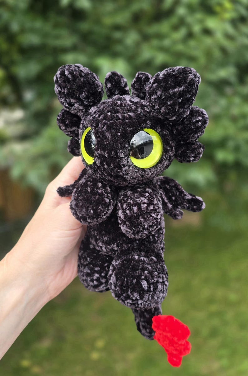 Puede incluir: Un drag&oacute;n de peluche negro hecho a mano con grandes ojos amarillos y una cola roja. El drag&oacute;n tiene peque&ntilde;as alas y rasgos parecidos a orejas en la cabeza. El peluche est&aacute; hecho de un material suave y texturizado.