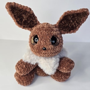 May include: A brown crochet plush toy resembling a fox-like creature with large, dark, glittery eyes, a small black nose, and a fluffy white collar around its neck. The toy features tall, pointed ears and a soft, textured yarn construction.