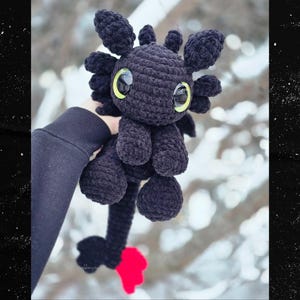 Puede incluir: Un peluche de drag&oacute;n negro hecho a mano con ganchillo, con ojos grandes, redondos y de color amarillo verdoso. El drag&oacute;n tiene una larga cola con punta roja y peque&ntilde;as alas redondeadas. El peluche se sostiene contra un fondo borroso y nevado.