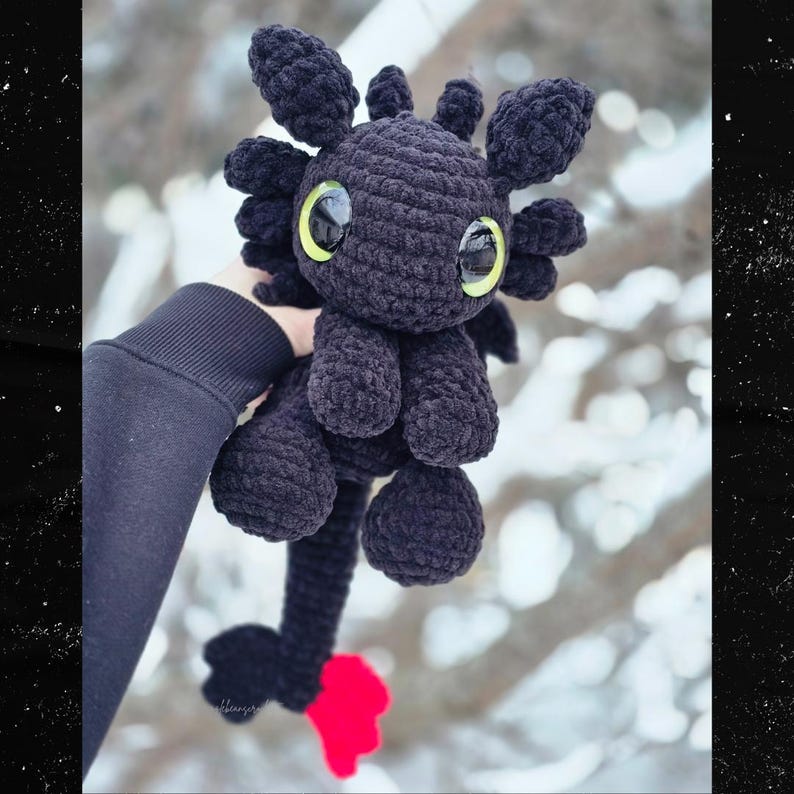 Puede incluir: Un peluche de drag&oacute;n negro hecho a mano con ganchillo, con ojos grandes, redondos y de color amarillo verdoso. El drag&oacute;n tiene una larga cola con punta roja y peque&ntilde;as alas redondeadas. El peluche se sostiene contra un fondo borroso y nevado.