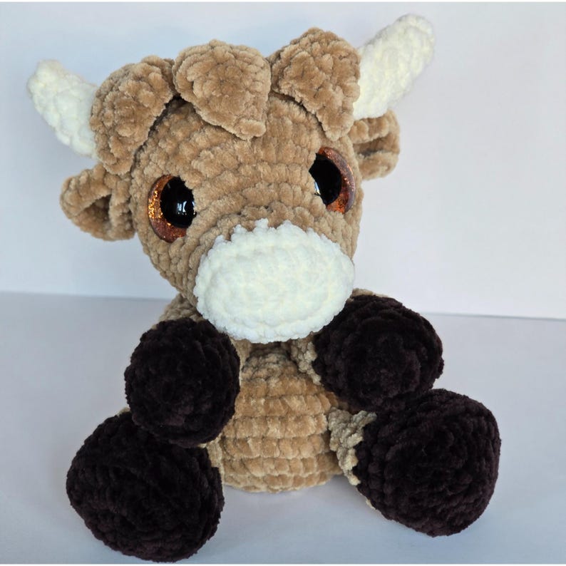 May include: A handmade Highland cow plush toy. The cow is mainly brown with white horns and a white muzzle. It has large, sparkly eyes and dark brown hooves. The toy is made of soft, textured yarn.