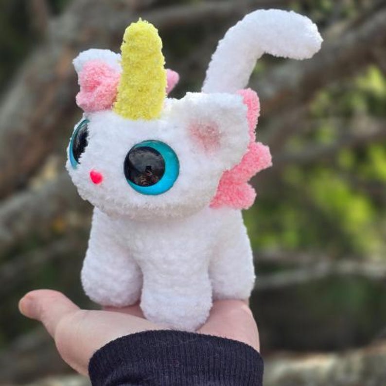May include: A white crocheted unicorn plush toy with a yellow horn, pink ears, and large blue eyes. The toy is sitting in a person's hand.