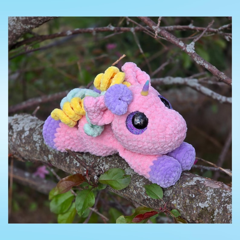 Peut inclure: Une peluche licorne rose au crochet, avec des sabots et des accents violets, une corne jaune et une crini&egrave;re multicolore. La licorne a de grands yeux scintillants et est pos&eacute;e sur une branche d'arbre. L'arri&egrave;re-plan est flou avec du feuillage vert.