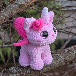 May include: A pink and white crocheted unicorn toy with a sparkly eye and a pink horn. The unicorn is sitting on a branch.