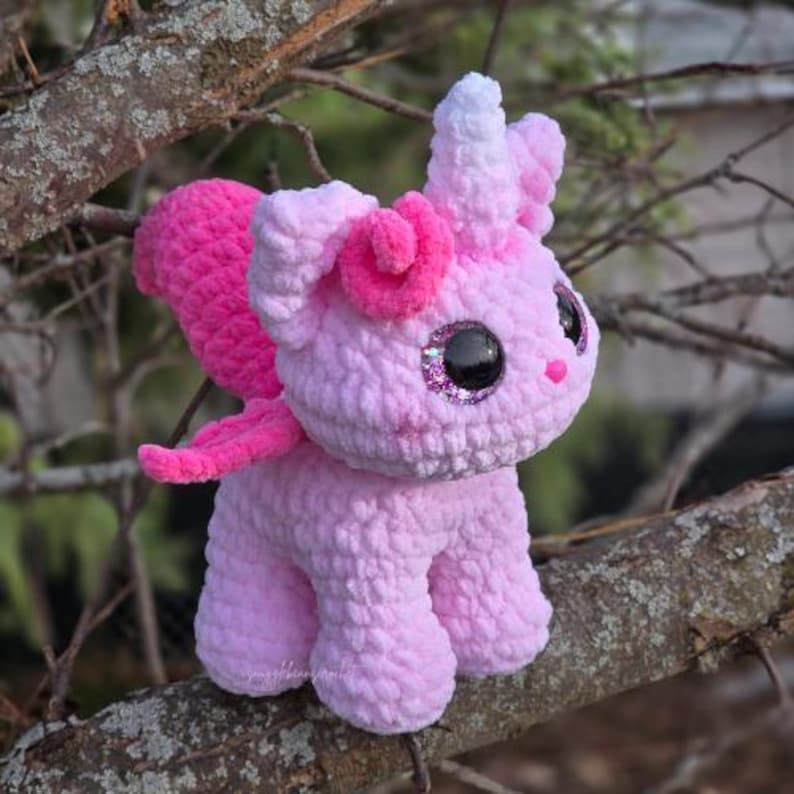 May include: A pink and white crocheted unicorn toy with a sparkly eye and a pink horn. The unicorn is sitting on a branch.